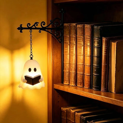A ghost-shaped wall lamp glowing warm white, holding a small book and hanging from a black metal bracket next to a wooden bookshelf filled with old books.